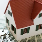 Model house with red roof sitting on banknotes next to a set of keys